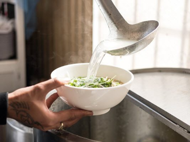 Một nồi nước dùng phở đậm đà đang được chan vào bát phở đã đầy đủ vắt, rau, thịt,... bằng một cái vá lớn. Khói bay nghi ngút từ nước dùng trong suốt, tạo cảm giác ấm áp và hấp dẫn.