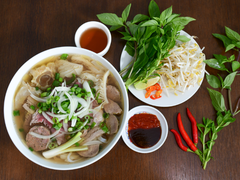 Tô phở bò đầy đặn với thịt bò tái, bò viên, gân, hành tây, hành lá, và măng, được dọn kèm đĩa rau sống gồm húng quế, giá đỗ, lát ớt, chanh và nước chấm. Bên cạnh là chén tương ớt – tương đen và vài trái ớt đỏ, tất cả bày biện trên mặt bàn gỗ nâu sẫm.