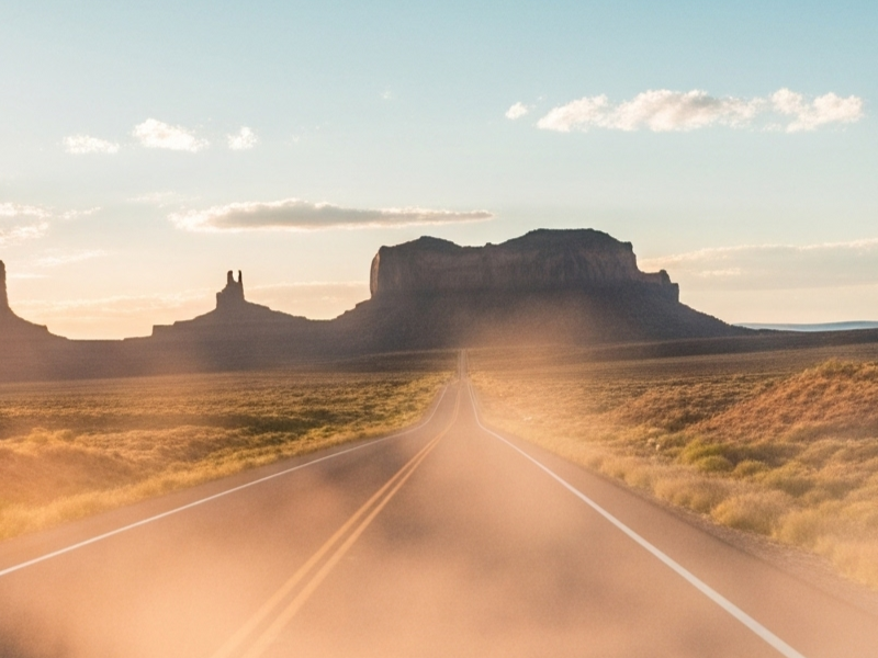 Hình ảnh mô tả một hành trình road trip tại Mỹ với con đường dài hun hút chạy xuyên qua vùng sa mạc rộng lớn, hai bên là núi đá hùng vĩ và bầu trời xanh ngắt, gợi cảm giác tự do, phiêu lưu và khám phá không giới hạn.