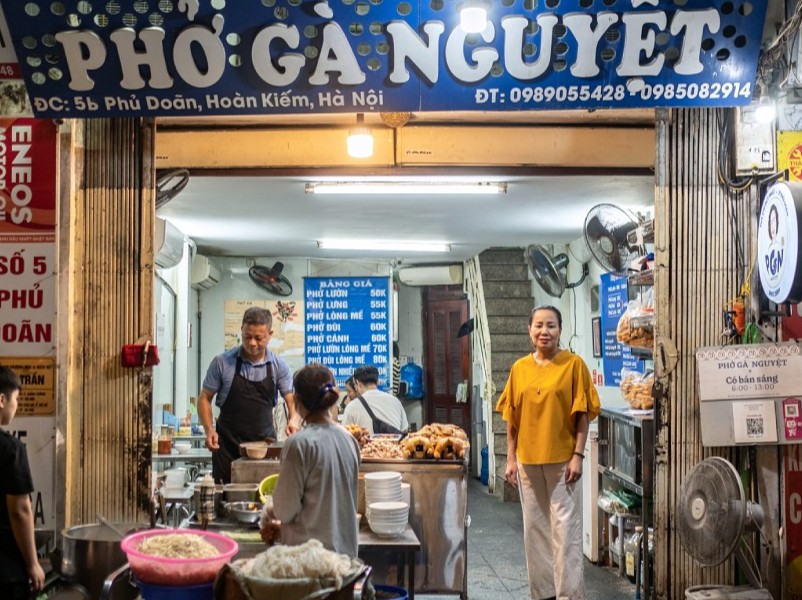 Mặt tiền quán Phở Gà Nguyệt tại số 5B Phủ Doãn, Hoàn Kiếm, Hà Nội với biển hiệu xanh nổi bật và thực đơn treo bên trong. Nhân viên đang chuẩn bị món ăn tại quầy bếp, phía trước có một người phụ nữ đứng cười thân thiện. Bên cạnh là bàn quầy với bánh quẩy và bát tô lớn, tạo nên không khí ấm cúng và nhộn nhịp của quán ăn sáng nổi tiếng.