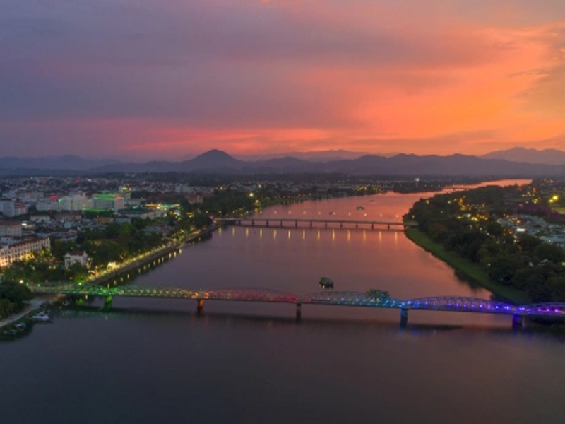 Du thuyền rồng với ánh đèn lung linh trôi trên sông Hương vào lúc hoàng hôn rực rỡ, phía xa là cầu Trường Tiền nổi bật dưới bầu trời đỏ cam tím huyền ảo của thành phố Huế.