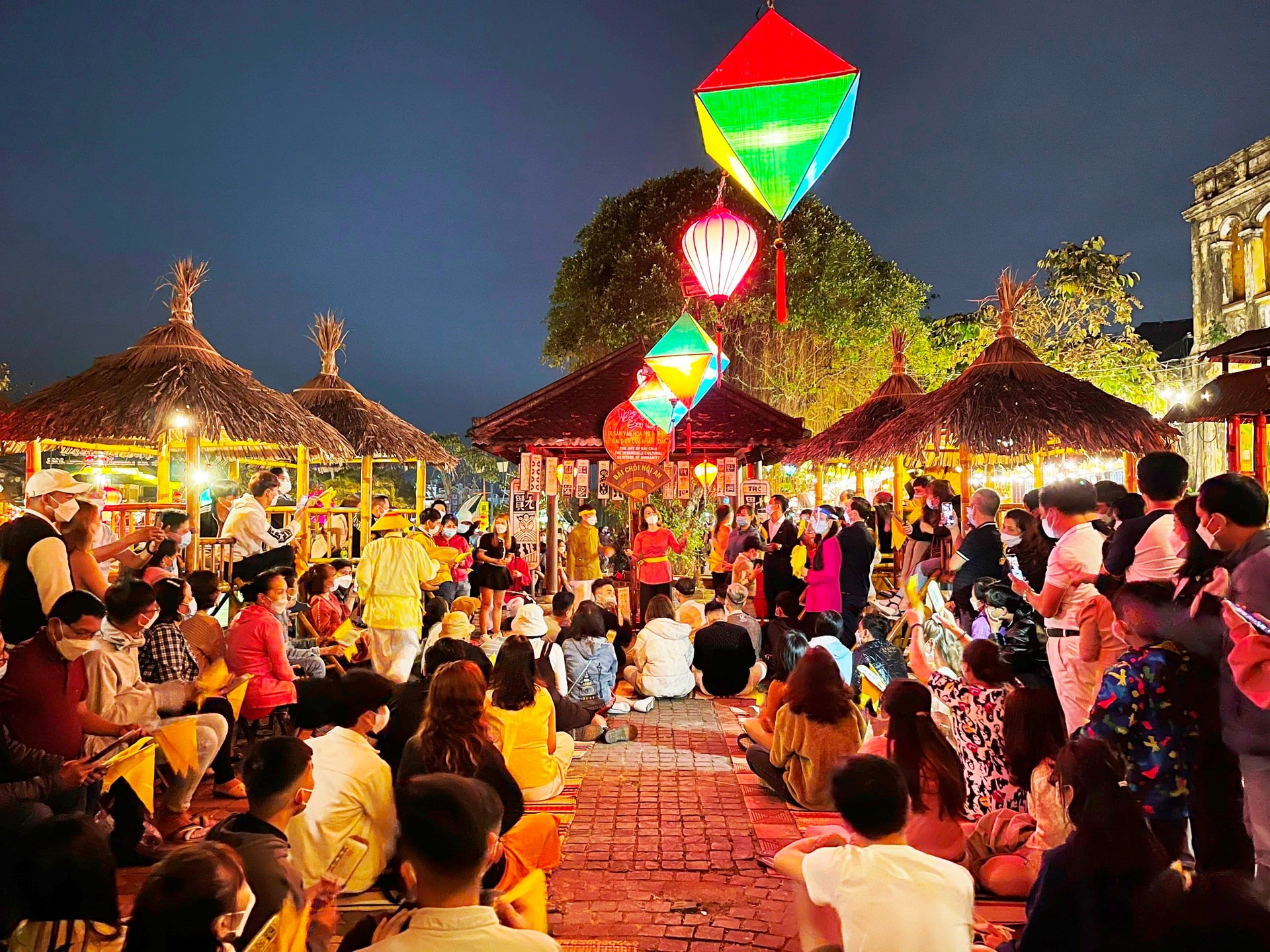 Người dân và du khách tham gia lễ hội văn hóa đường phố tại Việt Nam vào ban đêm, không gian rực rỡ với lồng đèn, nhà tranh và các tiết mục truyền thống